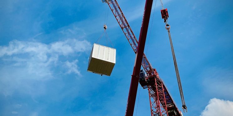 A red cane lifting a square pod made of wooden panels with a blue sky in the background